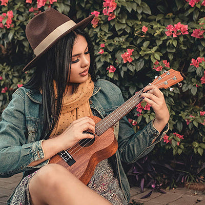Junge Frau mit Hut vor blühendem Busch spielt Ukulele