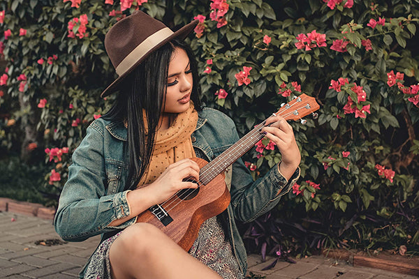 Junge Frau mit Hut vor blühendem Busch spielt Ukulele